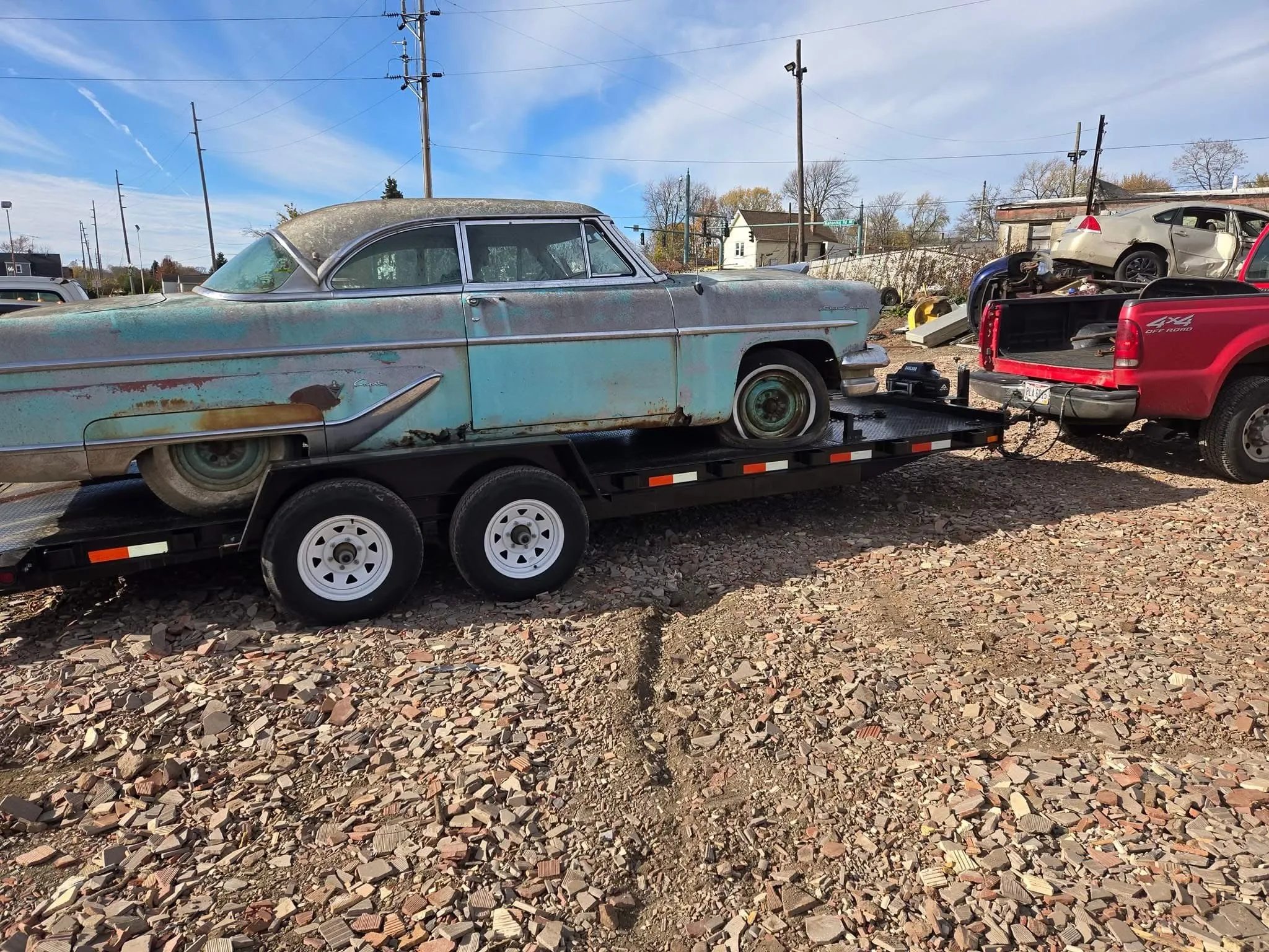 An old car, reminiscent of Ohio junk cars, is being transported on a car trailer across a dirt-covered area under a clear blue sky.